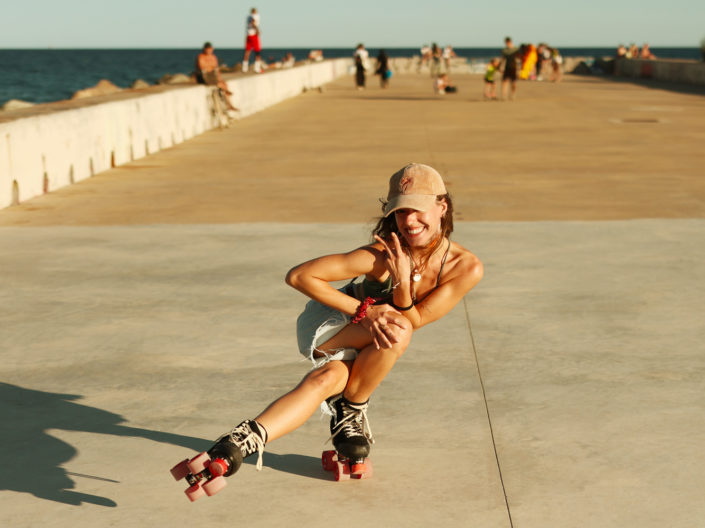 A joyful roller skater performing a move on a sunny pier, wearing a cap and smiling brightly, with the ocean and people in the background