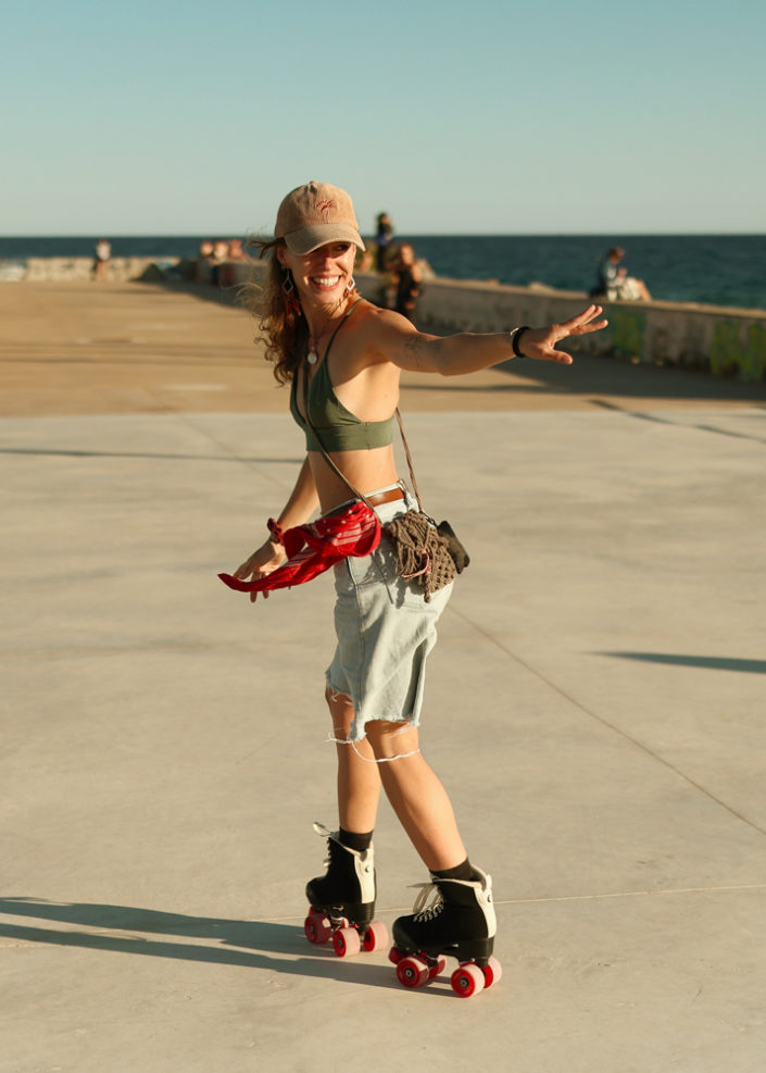 A joyful roller skater performing a move on a sunny pier, wearing a cap and smiling brightly, with the ocean and people in the background