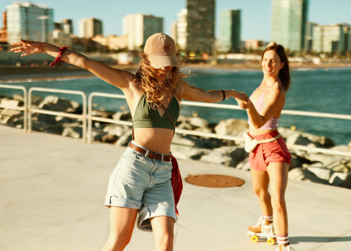 Two female roller skaters enjoying a sunny day on a pier, with one spinning joyfully while holding hands with the other, set against a cityscape and ocean backdrop.