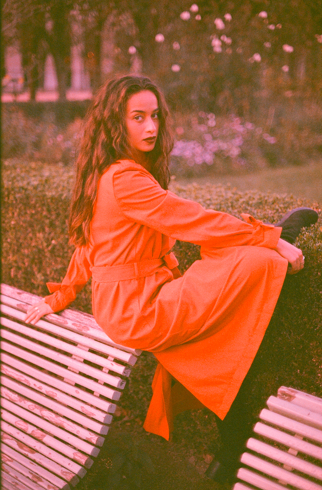 “Expired film photo of a woman wearing a red trench coat sitting in a park in Paris, taken with a Canon F1.”