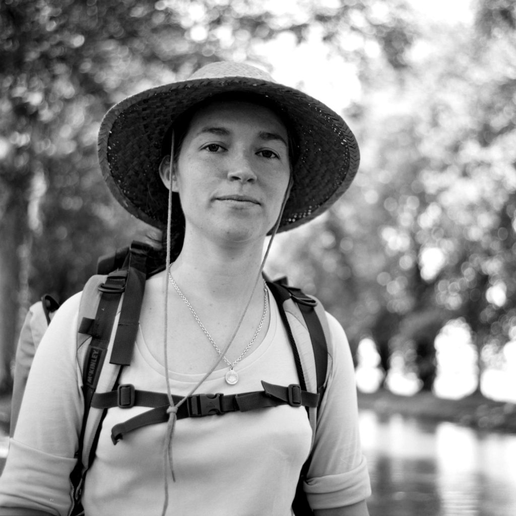 “Black and white portrait of a female pilgrim on the Saint-Jacques-de-Compostelle, wearing a backpack and hat, with nature visible in the background.”