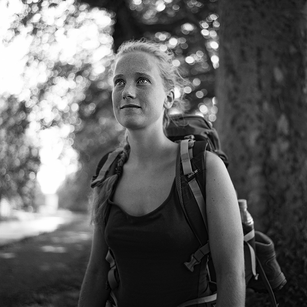 “Black and white portrait of a female pilgrim on the Saint-Jacques-de-Compostelle, wearing a backpack and hat, with nature visible in the background.”