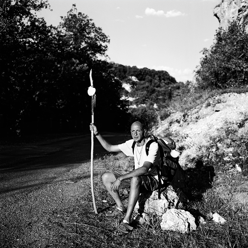 “Black and white portrait of a male pilgrim on the Saint-Jacques-de-Compostelle, wearing a backpack and hat, with nature visible in the background.”