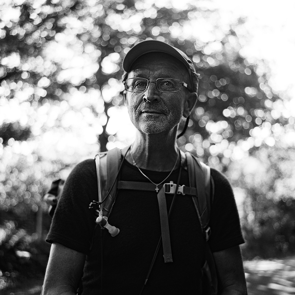 “Black and white portrait of a male pilgrim on the Saint-Jacques-de-Compostelle, wearing a backpack and hat, with nature visible in the background.”