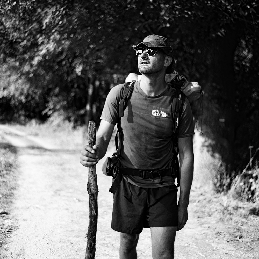 “Black and white portrait of a male pilgrim on the Saint-Jacques-de-Compostelle, wearing a backpack and hat, with nature visible in the background.”