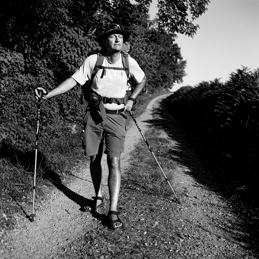 “Black and white portrait of a male pilgrim on the Saint-Jacques-de-Compostelle, wearing a backpack and hat, with nature visible in the background.”