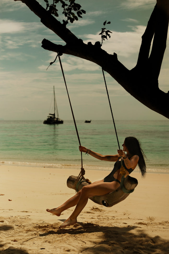 Agatha, an Italian woman, enjoying a rustic swing tied to a tree by the beach, with a sailboat floating in the background on calm turquoise waters of Thailand