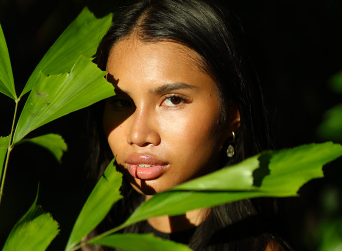 Portrait of a Thai woman in the jungle, framed by vibrant green palm leaves, with sunlight casting a soft glow on her face.