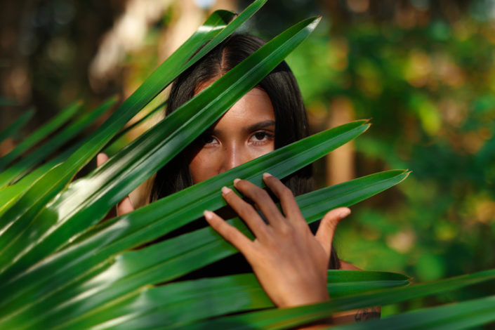 Portrait of a Thai woman in the jungle, framed by vibrant green palm leaves, with sunlight casting a soft glow on her face.