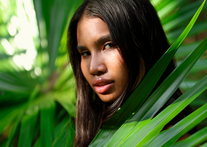 Portrait of a Thai woman in the jungle, framed by vibrant green palm leaves, with sunlight casting a soft glow on her face.