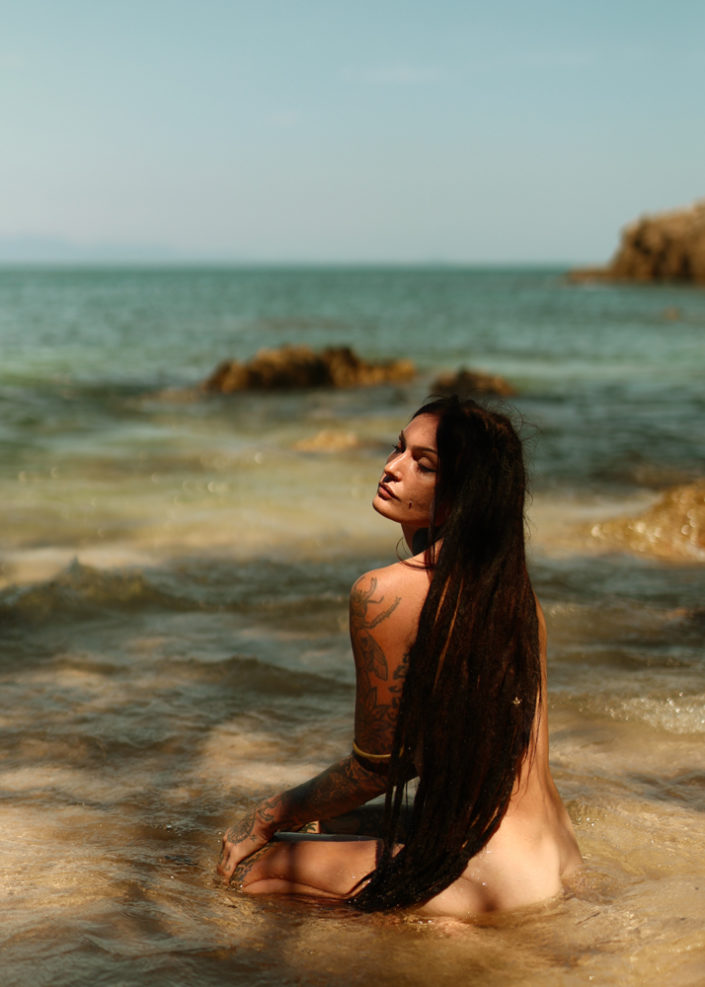 “Portrait of a woman with dreadlocks sitting in clear ocean water, surrounded by rocks under natural sunlight.”