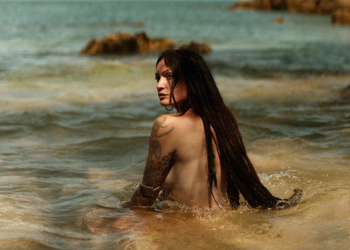 “Portrait of a woman with dreadlocks sitting in clear ocean water, surrounded by rocks under natural sunlight.”