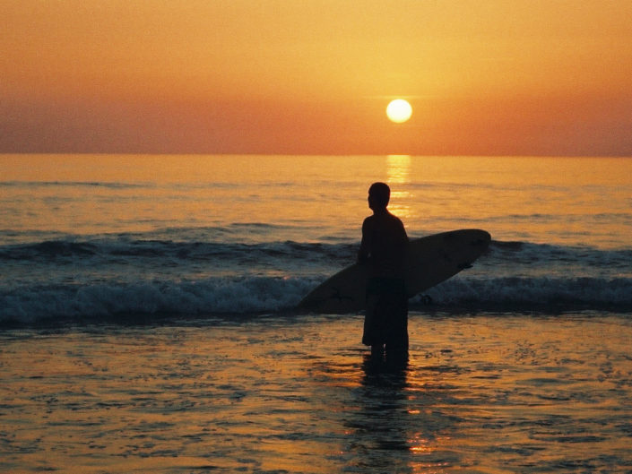 “Silhouette of a surfer holding a board at sunset, framed by vibrant orange, yellow, and red hues in the sky, with a landscape format emphasizing the serene beauty of the scene.”